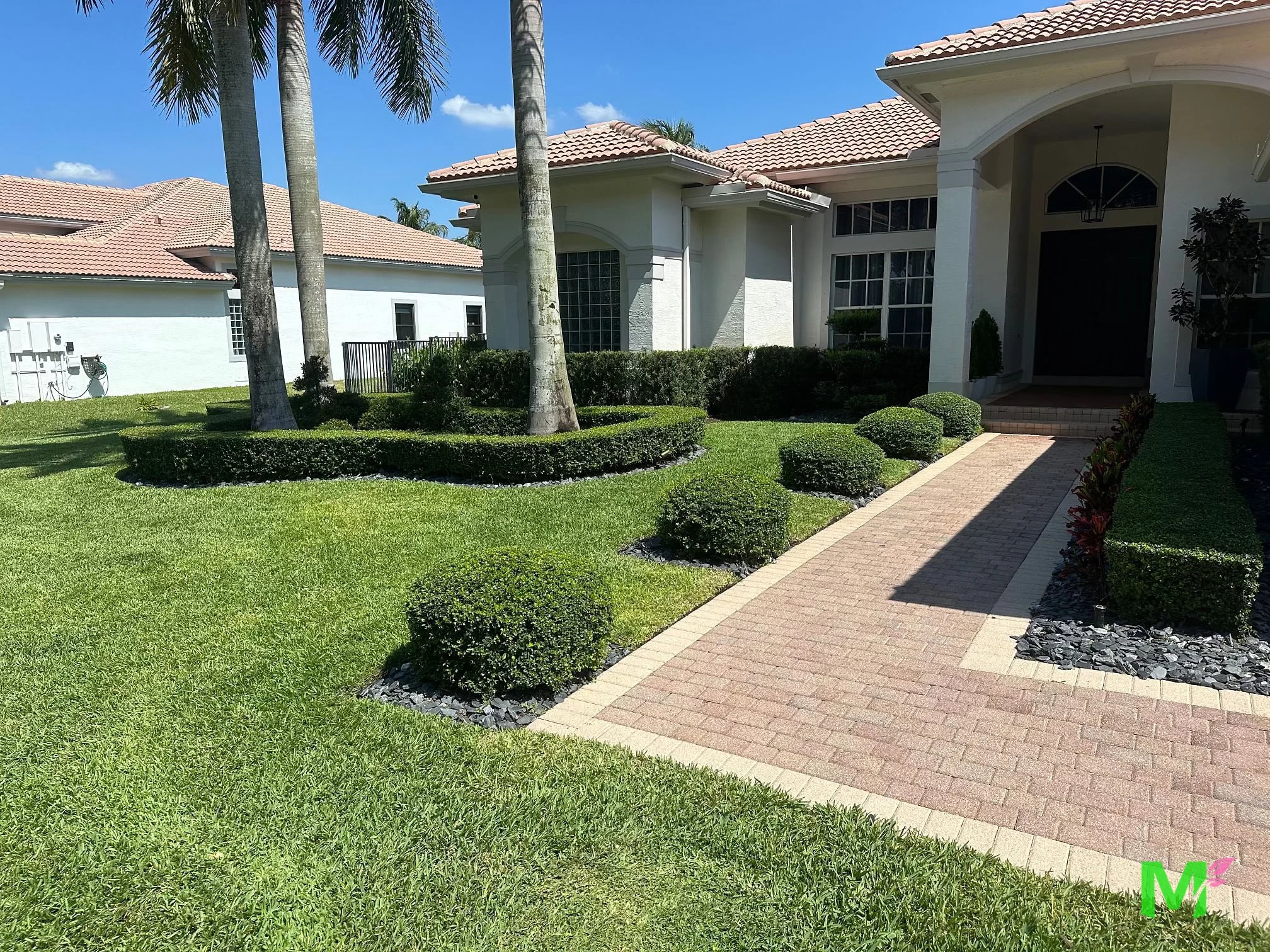 Brick paver walkway to a front entry, lined with rounded shrubs, clipped hedges and black river rock