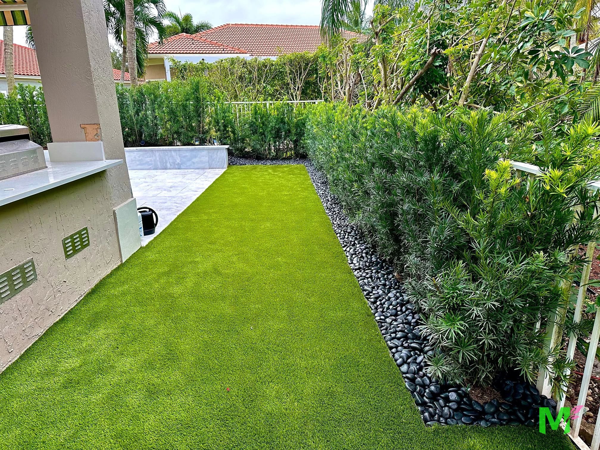 Patio trasero con césped artificial, un seto alto de privacidad y un borde de piedra de río negra