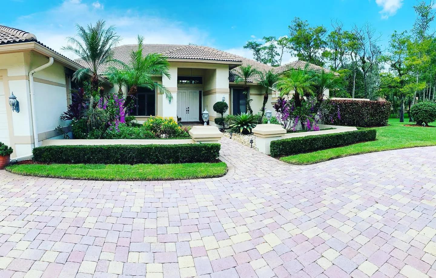 Fachada frontal de una residencia con plantas tropicales en capas, setos recortados y un camino de adoquines hasta la entrada