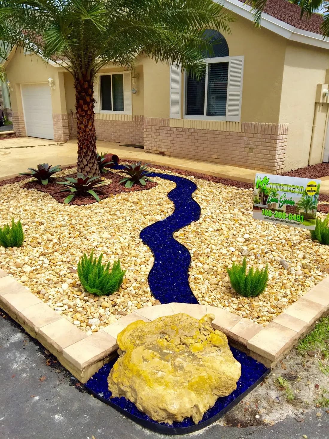 Cantero de xeriscape con un arroyo seco de vidrio azul cobalto, bromelias y un letrero de Montenegro Lawn Service