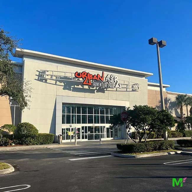 Entrada de una plaza comercial con setos cuidados y árboles de sombra